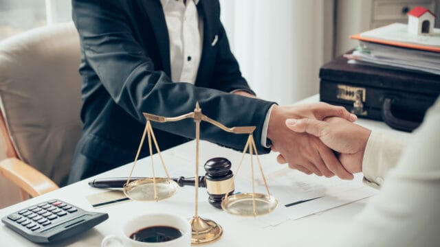 Two professionals shaking hands across a desk with a brass scale, calculator and contract papers