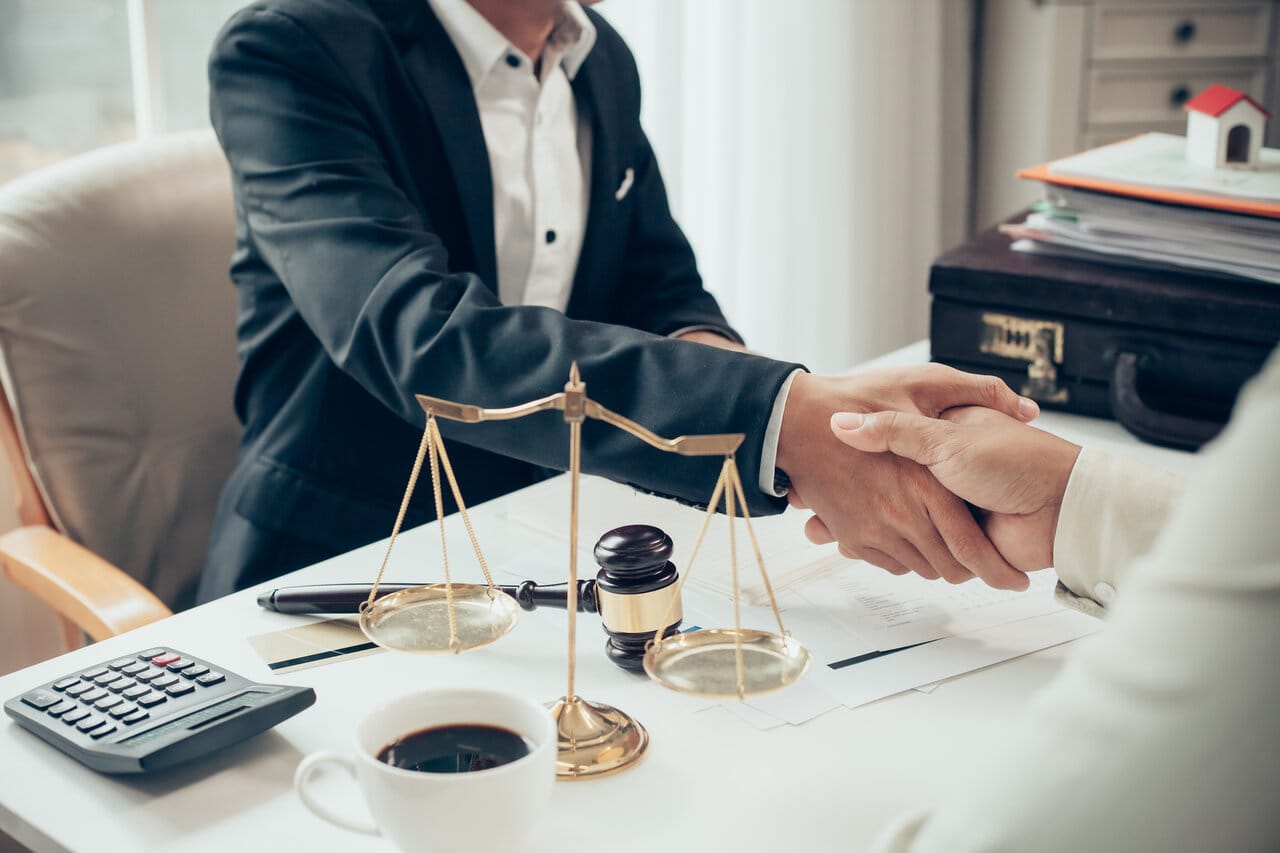 Two professionals shaking hands across a desk with a brass scale, calculator and contract papers