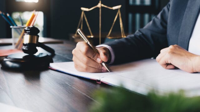 Hand writing notes at a desk with a brass scale and gavel in the foreground