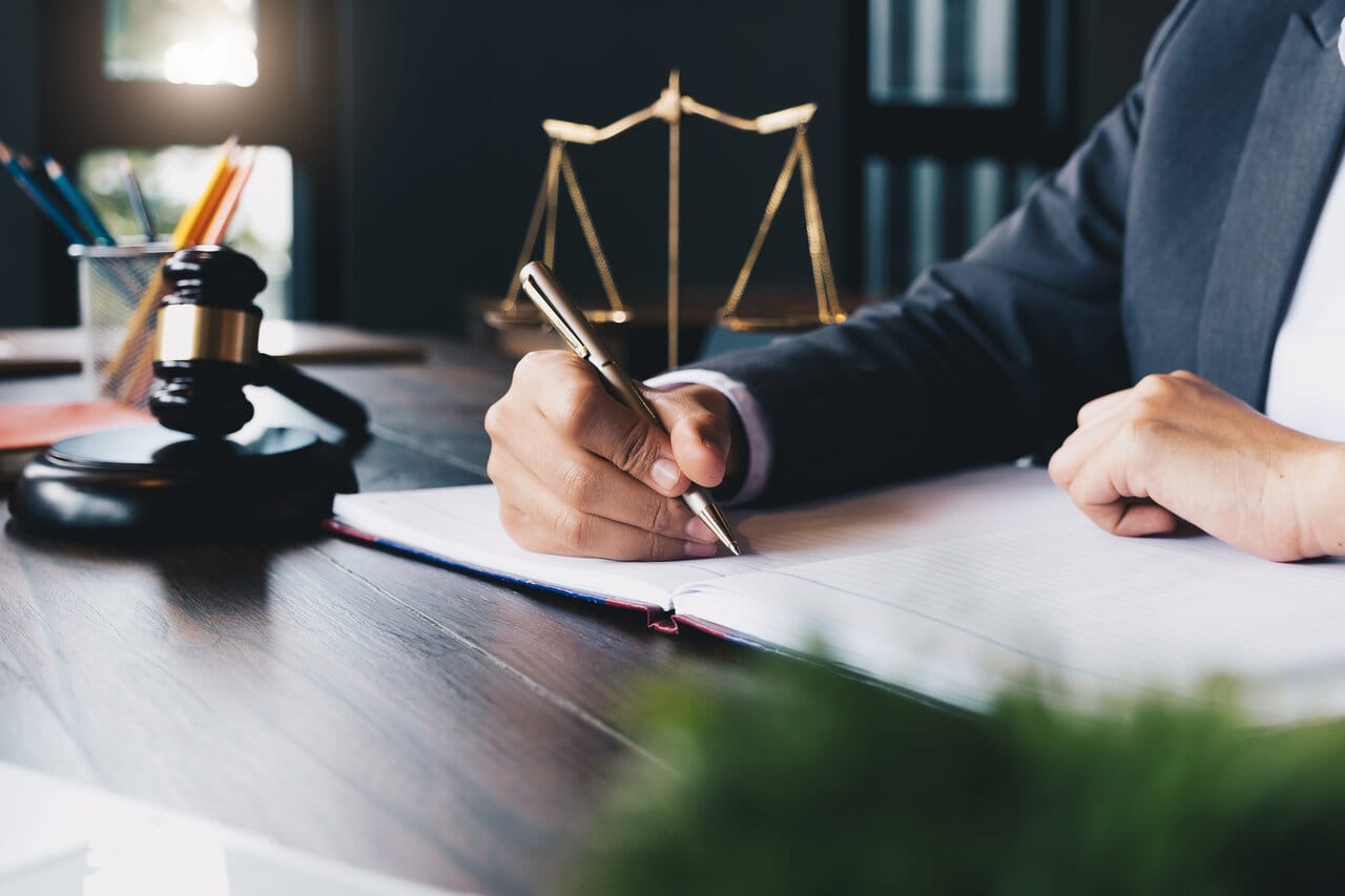 Hand writing notes at a desk with a brass scale and gavel in the foreground