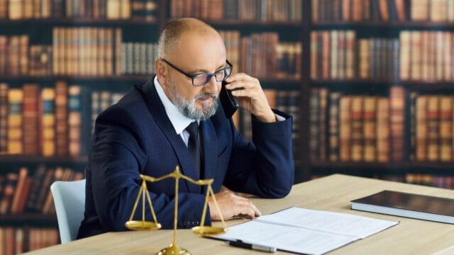Senior lawyer taking a phone call at his desk in front of a wall of law books, brass scale beside him