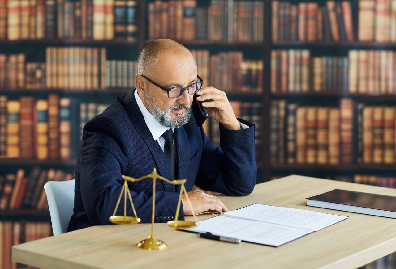 Senior lawyer taking a phone call at his desk in front of a wall of law books, brass scale beside him