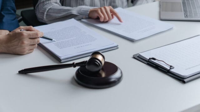 Hands reviewing stacked documents on clipboards beside a gavel and a laptop